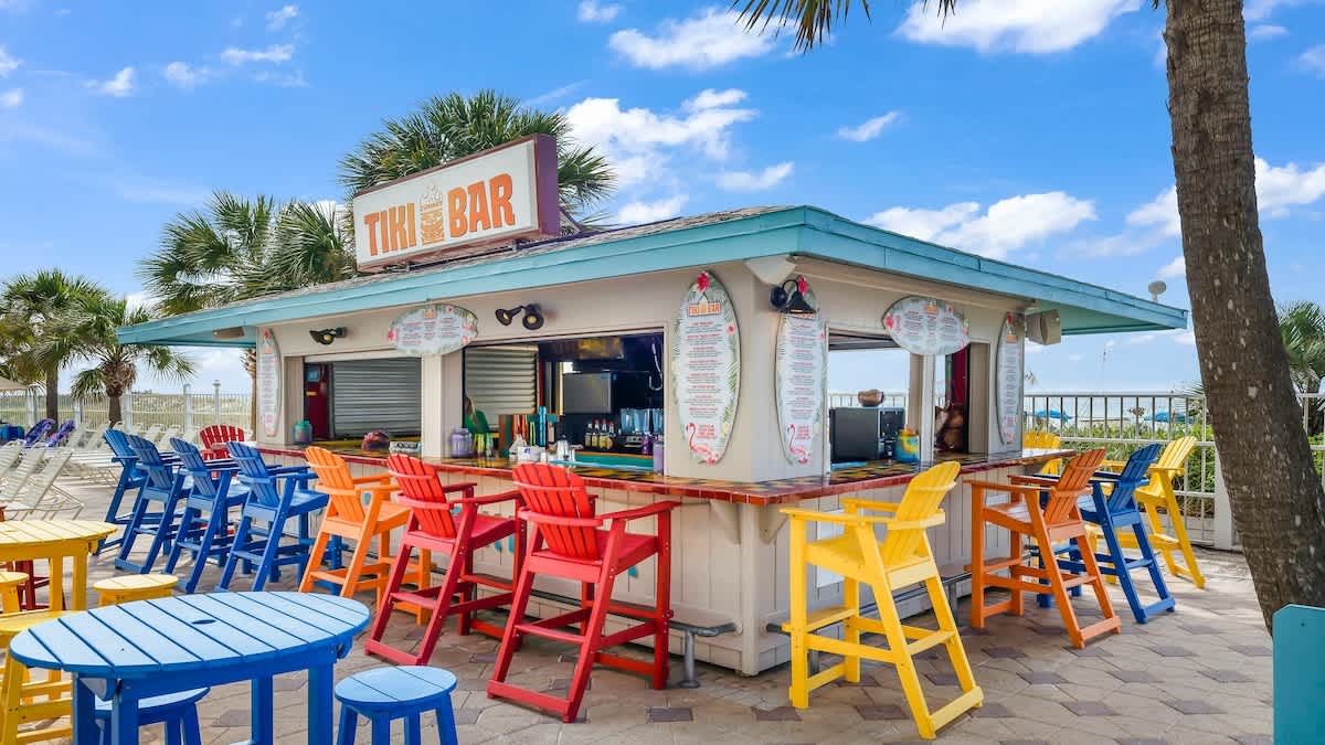 The Summit 1122  – Tiki bar with colorful chairs, overview of palm trees and beach in the background under blue sky.