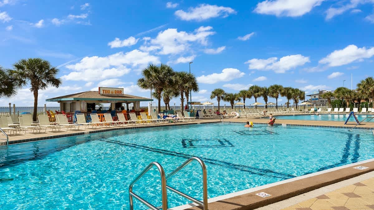 The Summit 1122  – Swimming pool area with a tiki bar, sun loungers, and palm trees against a blue sky.