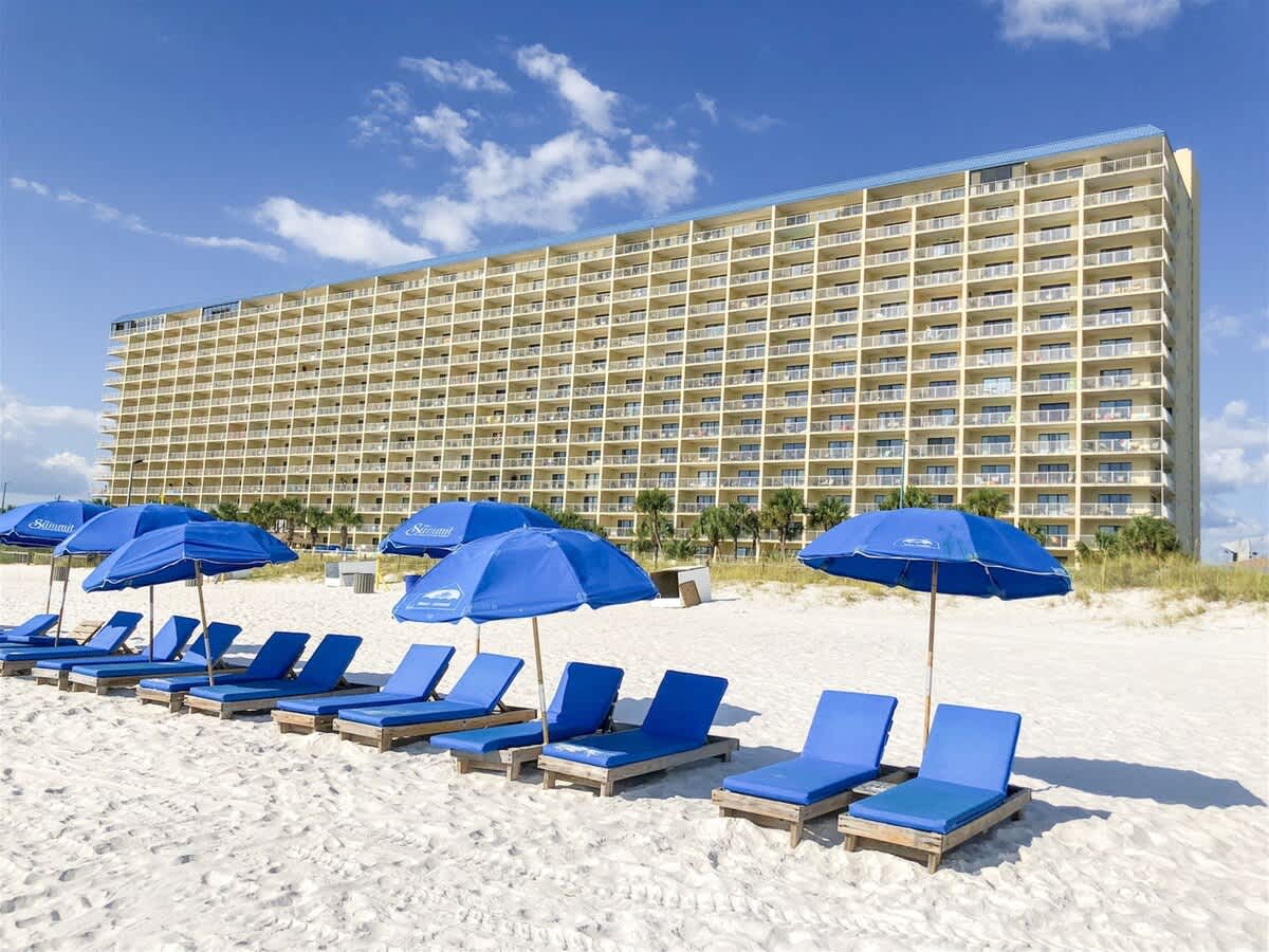 The Summit 618 – Beachfront view of a tall condominium with blue lounge chairs and umbrellas on white sand.