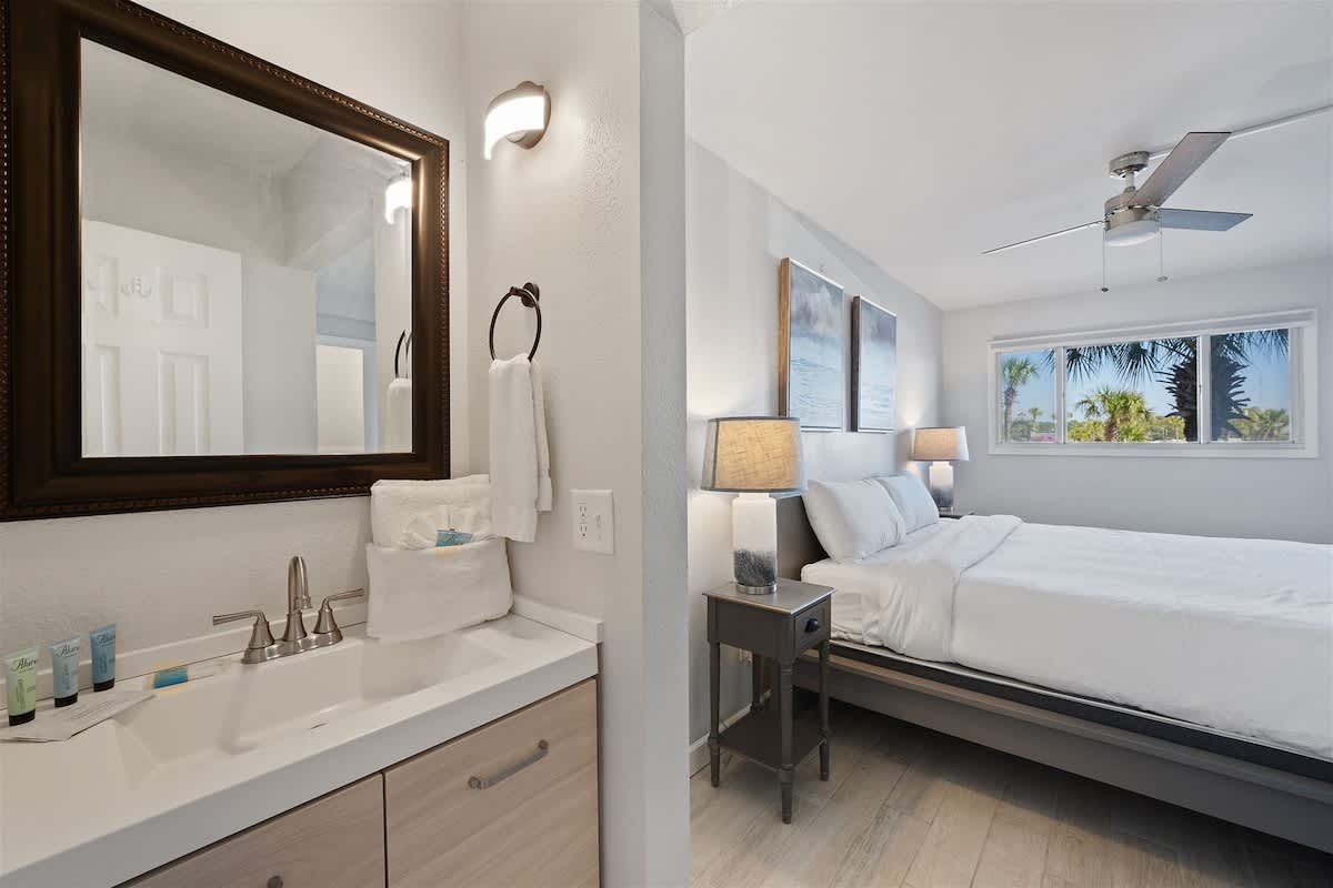 The Summit 220 – Bathroom with sink and mirror, adjacent bedroom featuring a bed, lamps, and a window with palm tree views.