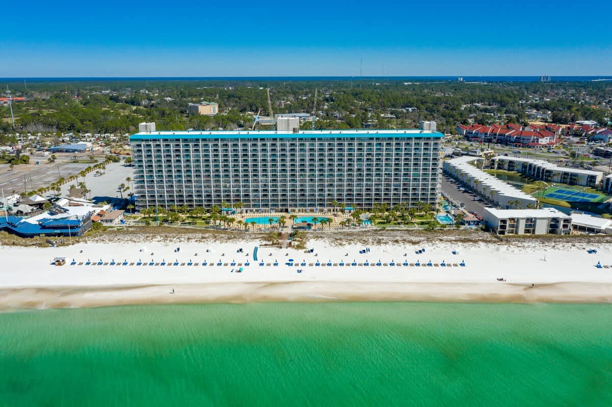 The Summit 602 – Aerial view of a large beachfront hotel with swimming pool and beach chairs along white sand.