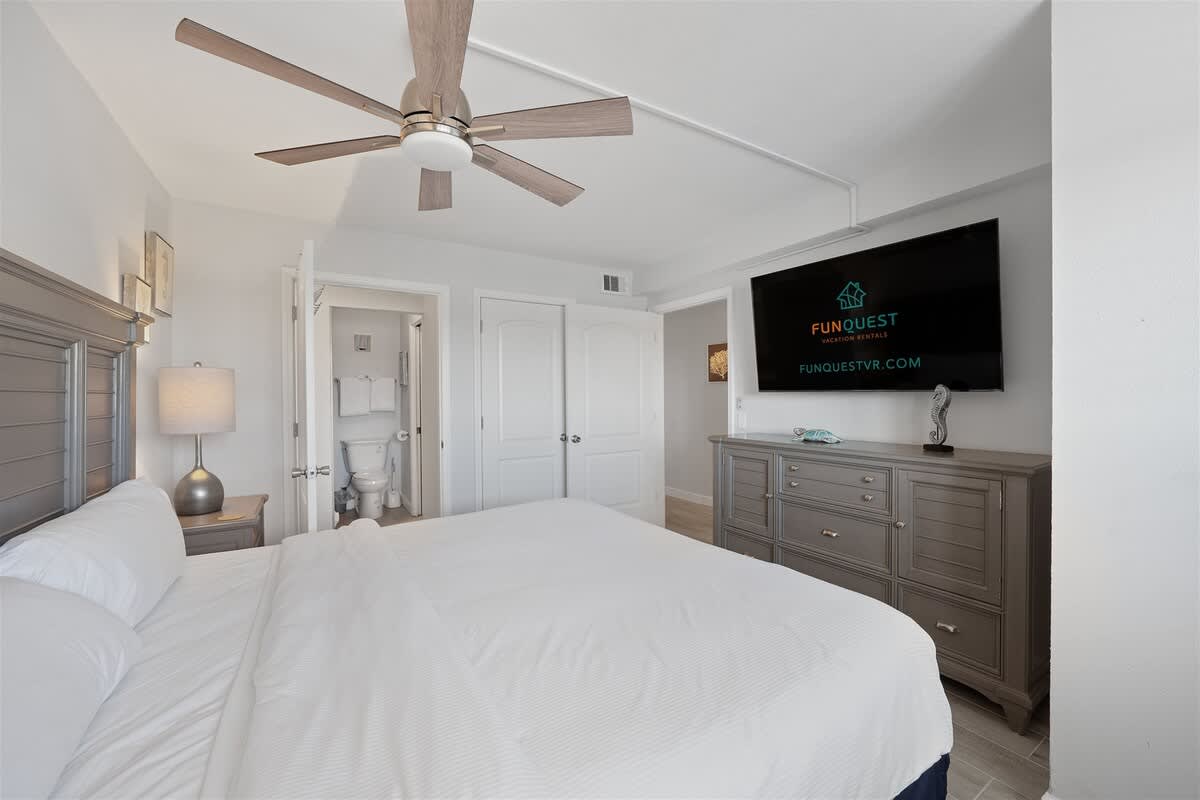 The Summit 621 – Bright bedroom with a king bed, ceiling fan, dresser, large TV, and en-suite bathroom door.