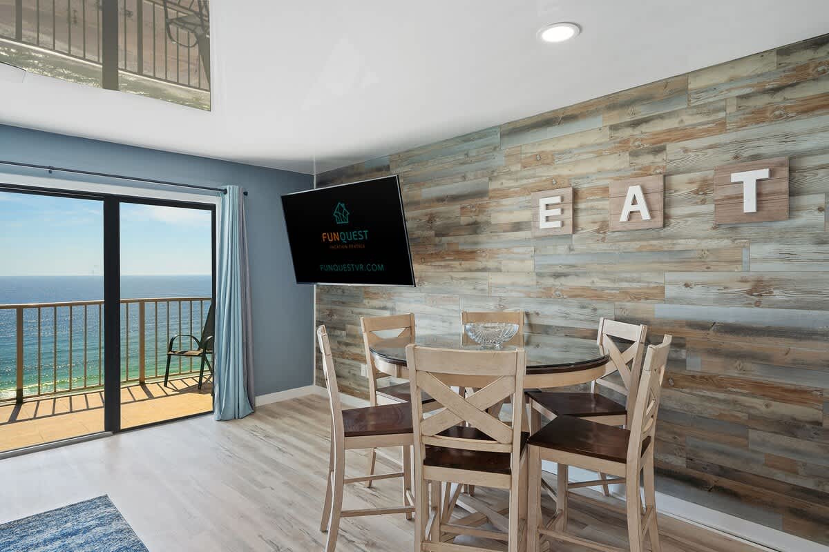 The Summit 1408 – Dining area with circular table, wooden chairs, wall-mounted TV, and ocean view through sliding glass doors.
