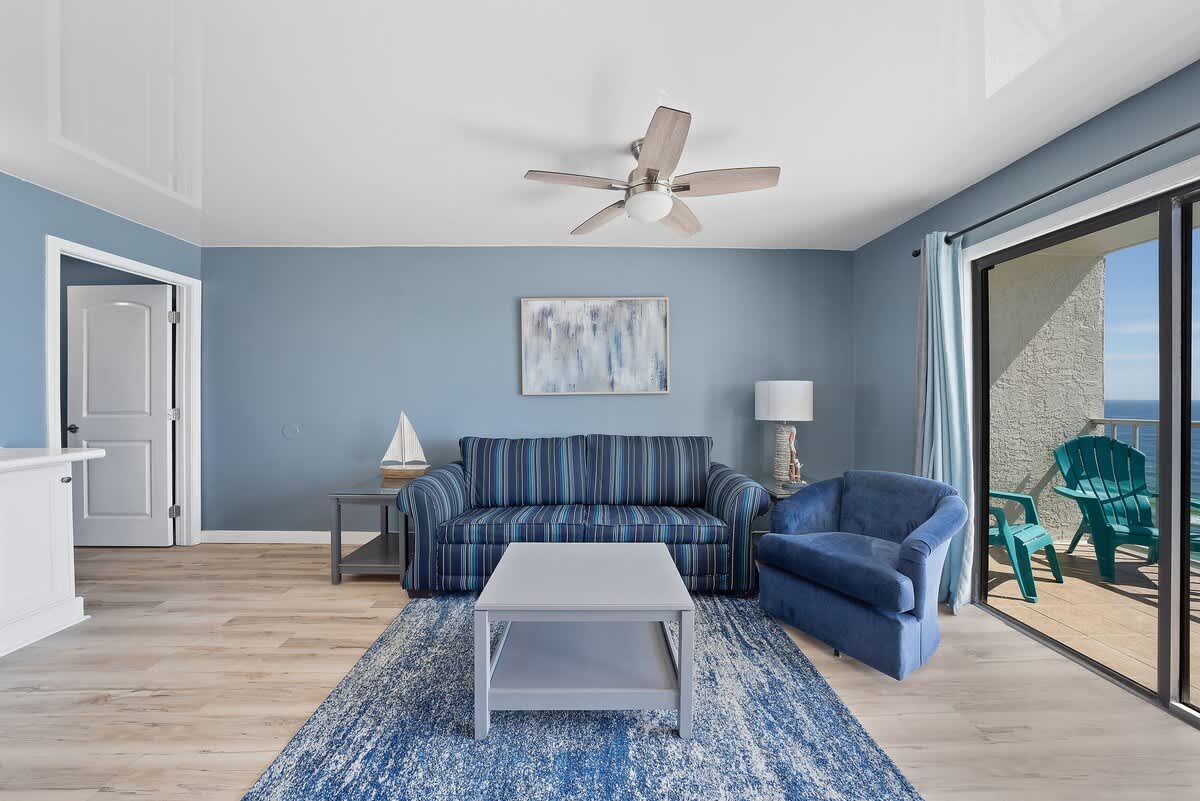 The Summit 1408 – Living room with blue walls, striped couch, table, and sliding doors to a balcony with sea view.