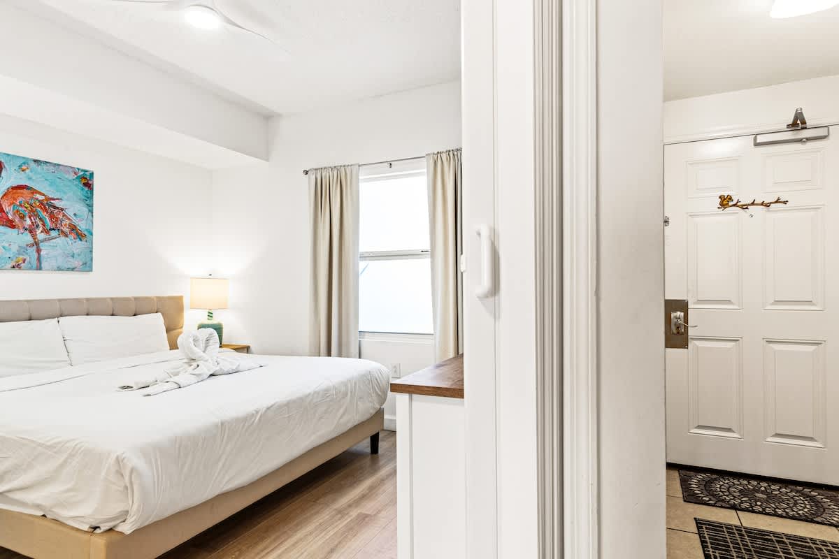 Seychelles 1006 – Bright bedroom with a large bed, lamp, and window, visible entrance door.
