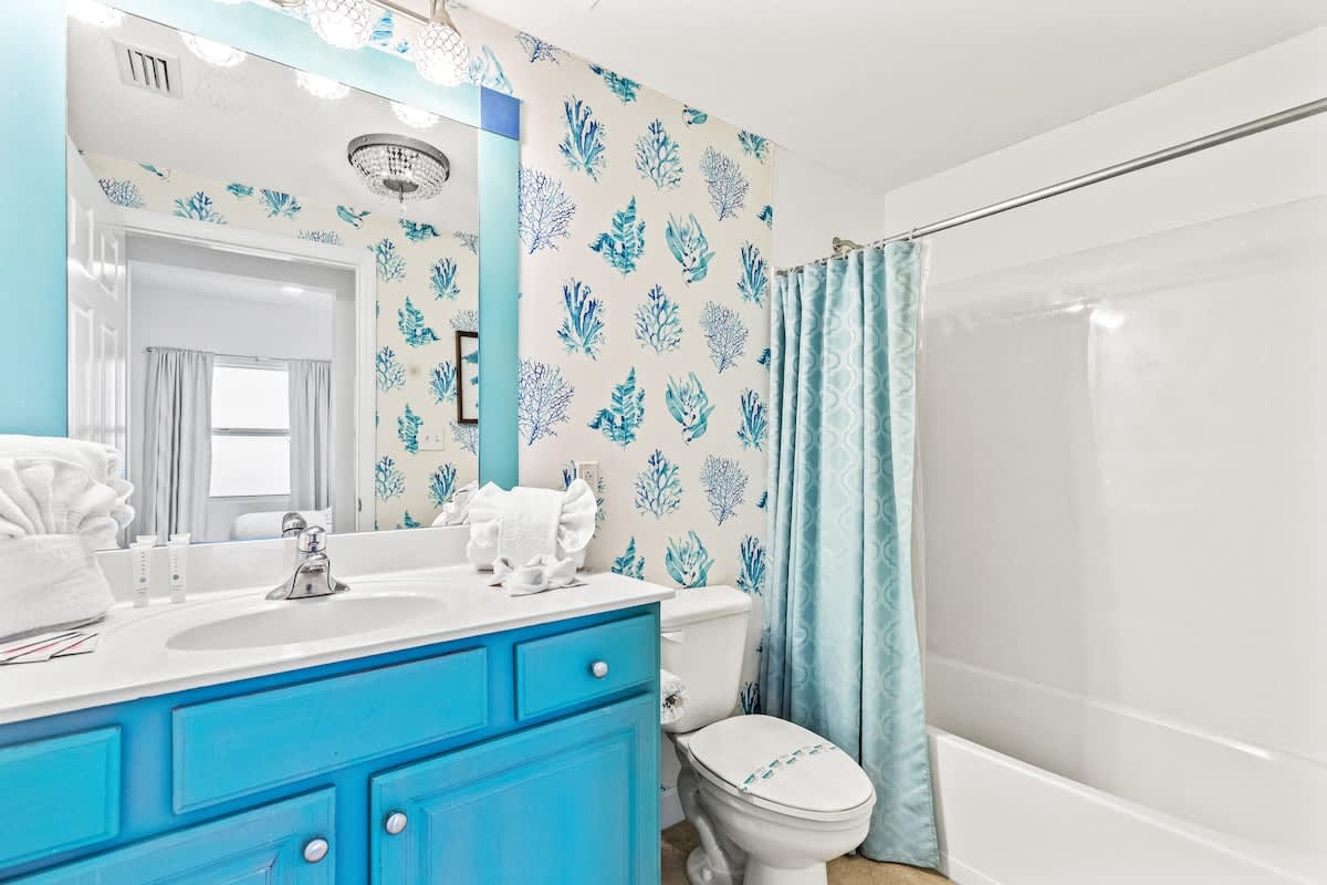 Seychelles 1006 – Bright bathroom with blue accents, coral-patterned wallpaper, and a bathtub with a shower curtain.