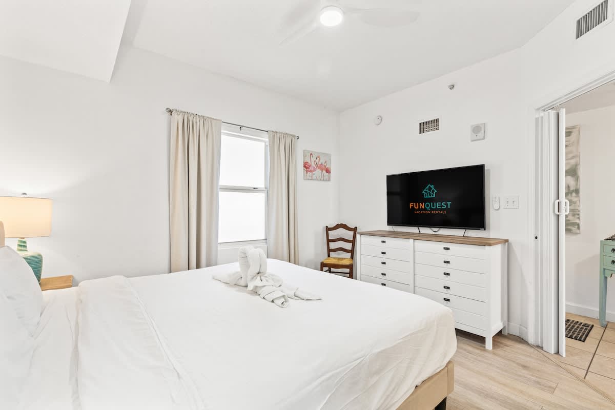 Seychelles 1006 – Bright bedroom with a king bed, dresser, TV, and window with curtains.