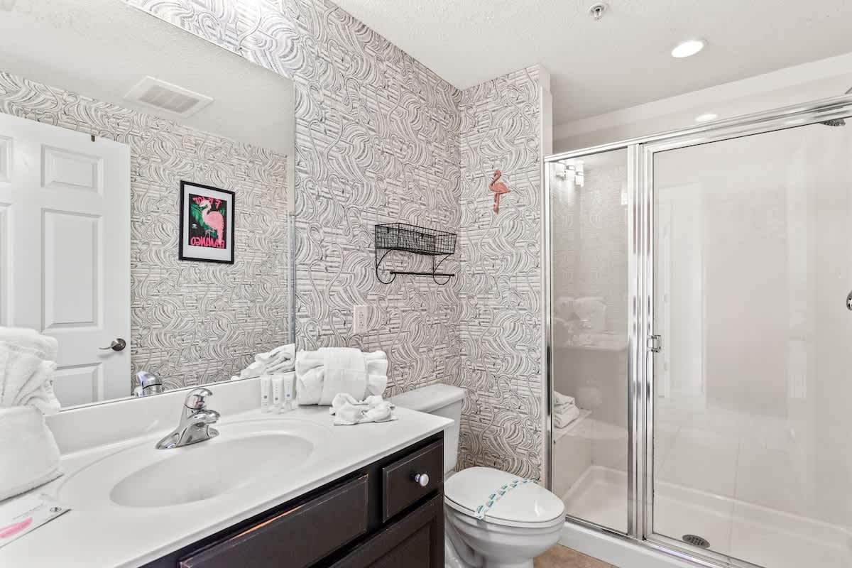 Seychelles 1006 – Modern bathroom with a double sink, shower, decorative wallpaper, and towels folded on the counter.