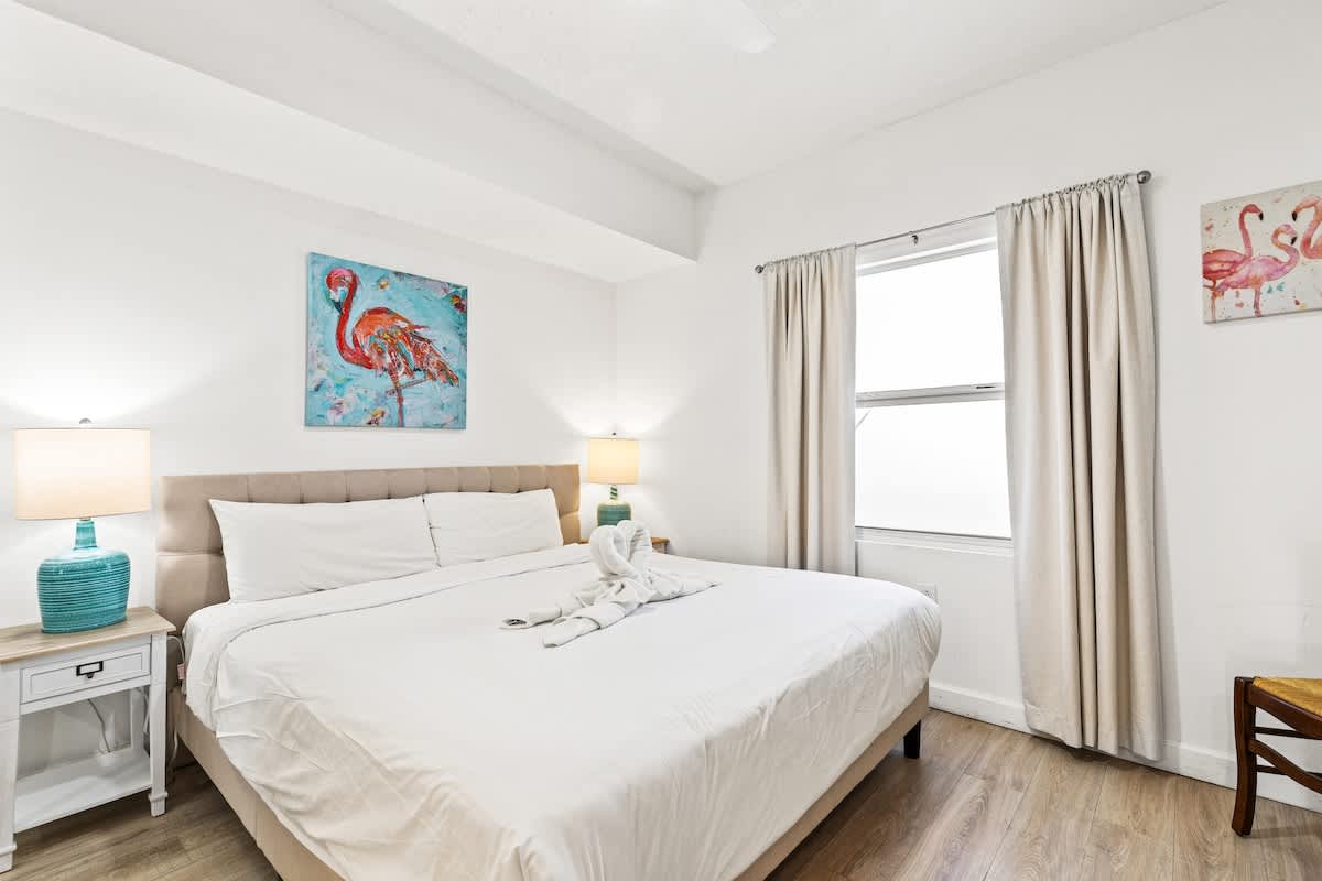 Seychelles 1006 – Bright bedroom with a large bed, nightstand, and window adorned with curtains.