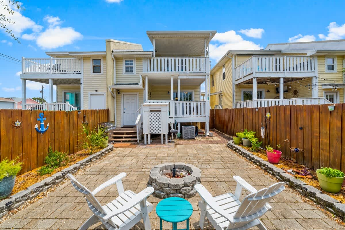 7405 Beach Dr A – Two white chairs and a fire pit in a paved courtyard with a wooden fence and surrounding plants.