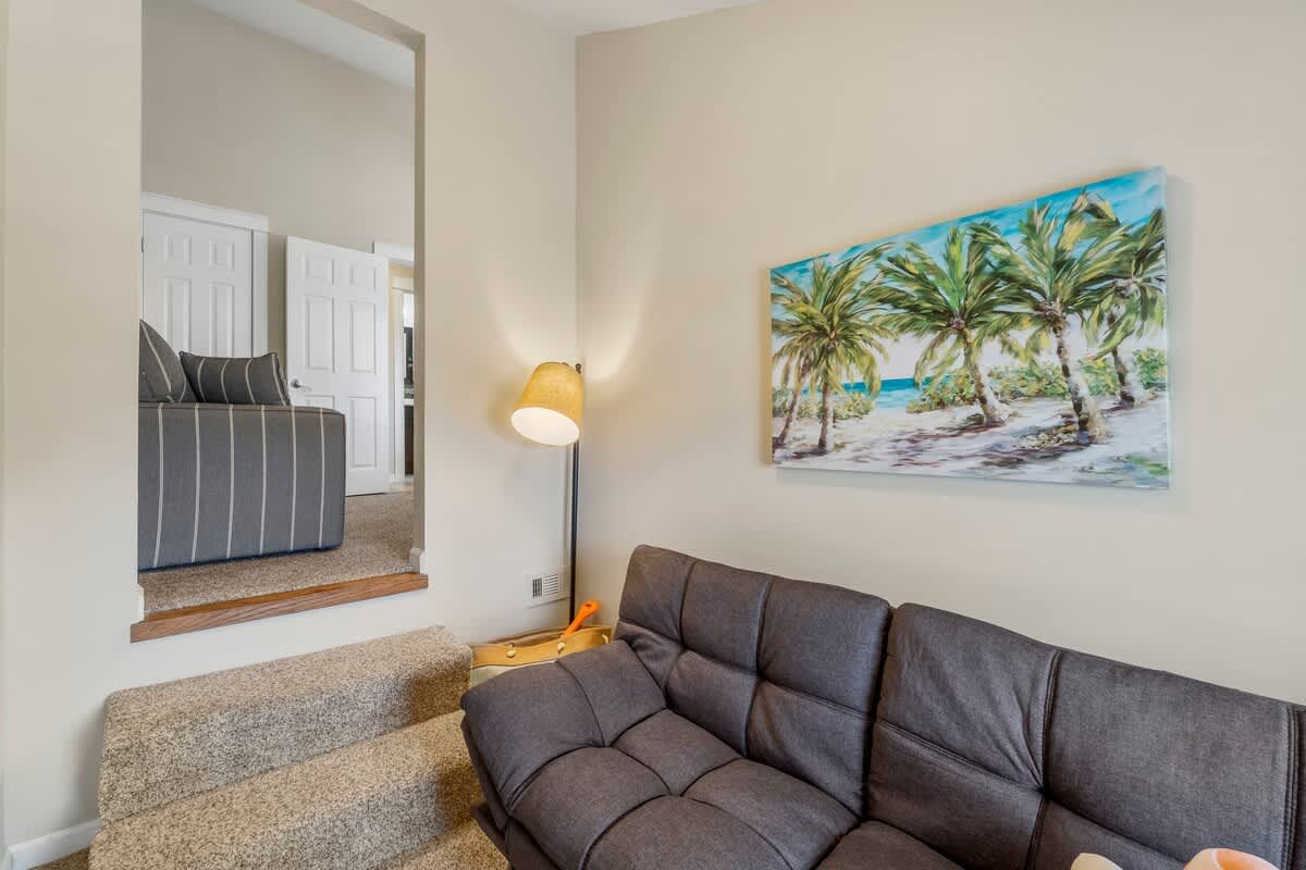 7405 Beach Dr A – Living room with grey sofa, lamp, and palm tree artwork, featuring steps to another room.