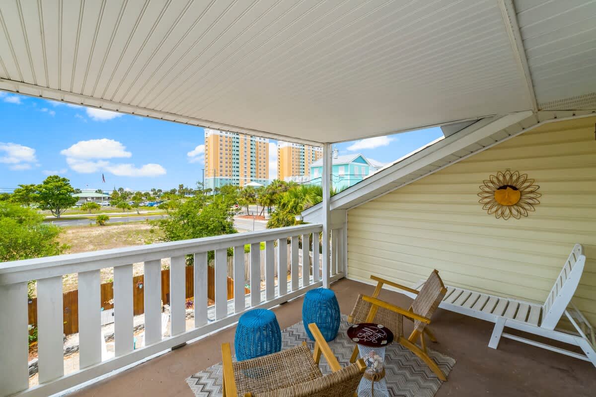 7405 Beach Dr A – Covered balcony with wicker chairs, blue accent stools, and a view of nearby buildings and greenery.