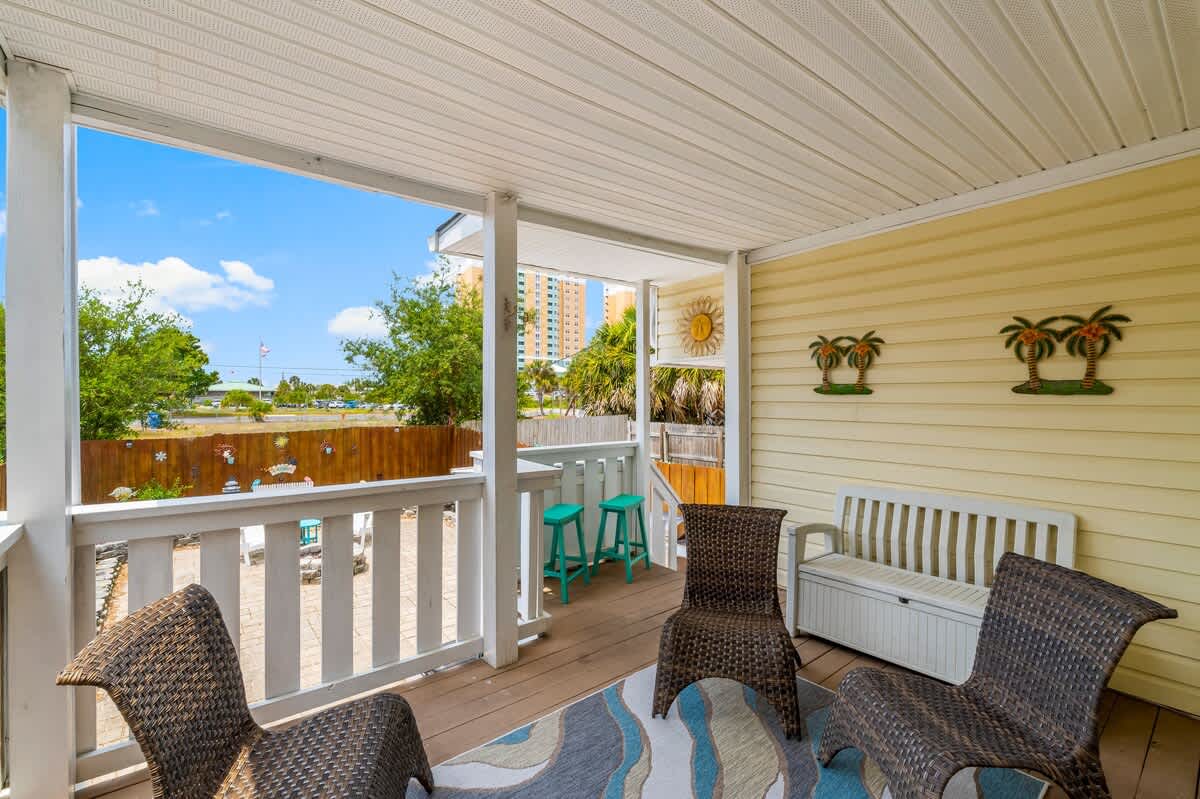 7405 Beach Dr A – Covered porch with wicker furniture, tropical decorations, and a view of greenery and a distant building.