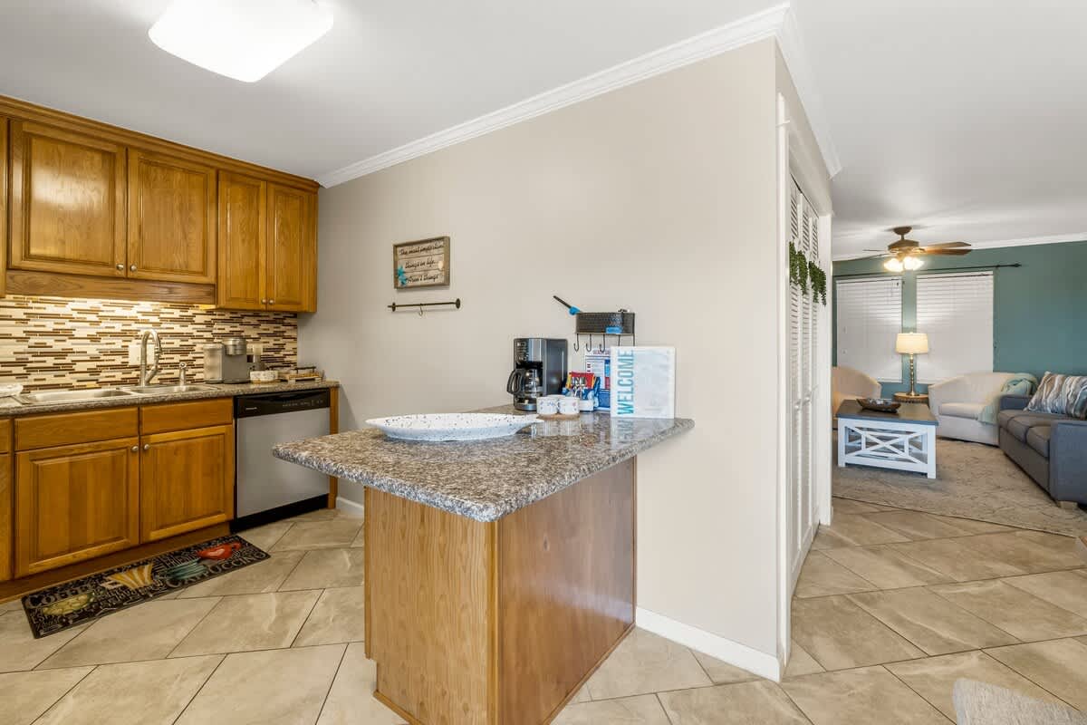 7405 Beach Dr A – Open kitchen with wooden cabinets and granite countertops, leading to a living area with sofa and light-colored chairs.