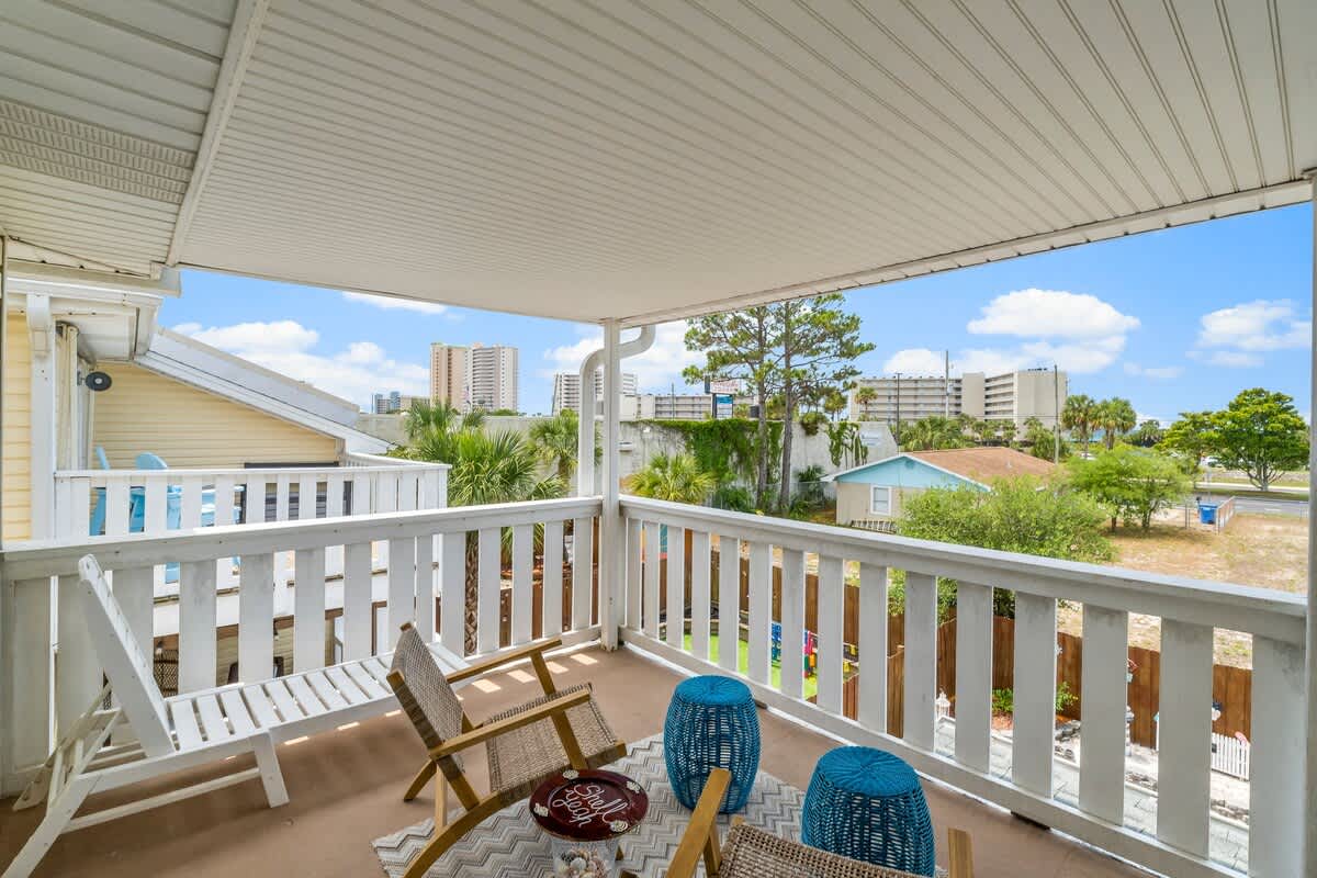 7405 Beach Dr A – Covered balcony with two chairs, blue tables, and views of nearby buildings and palm trees.