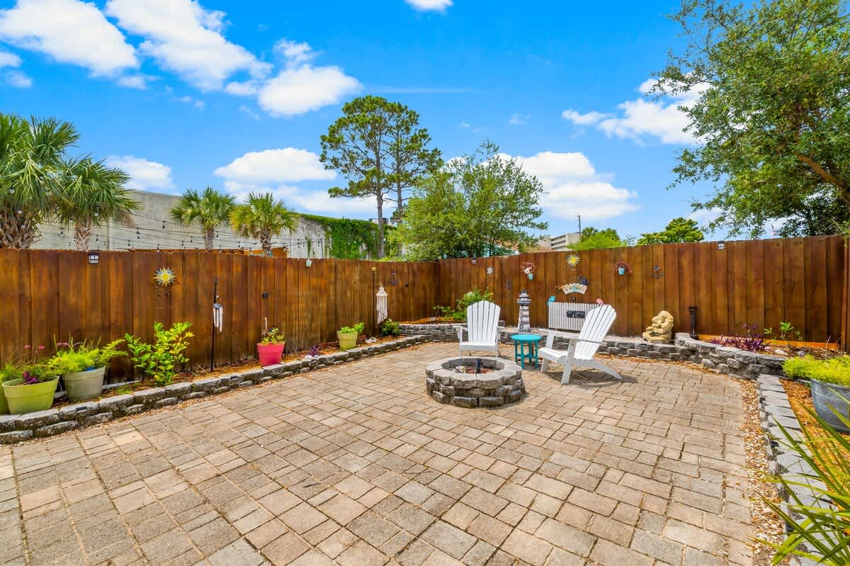 7405 Beach Dr A – Paved outdoor patio with two white chairs, fire pit, and various potted plants in a fenced yard.