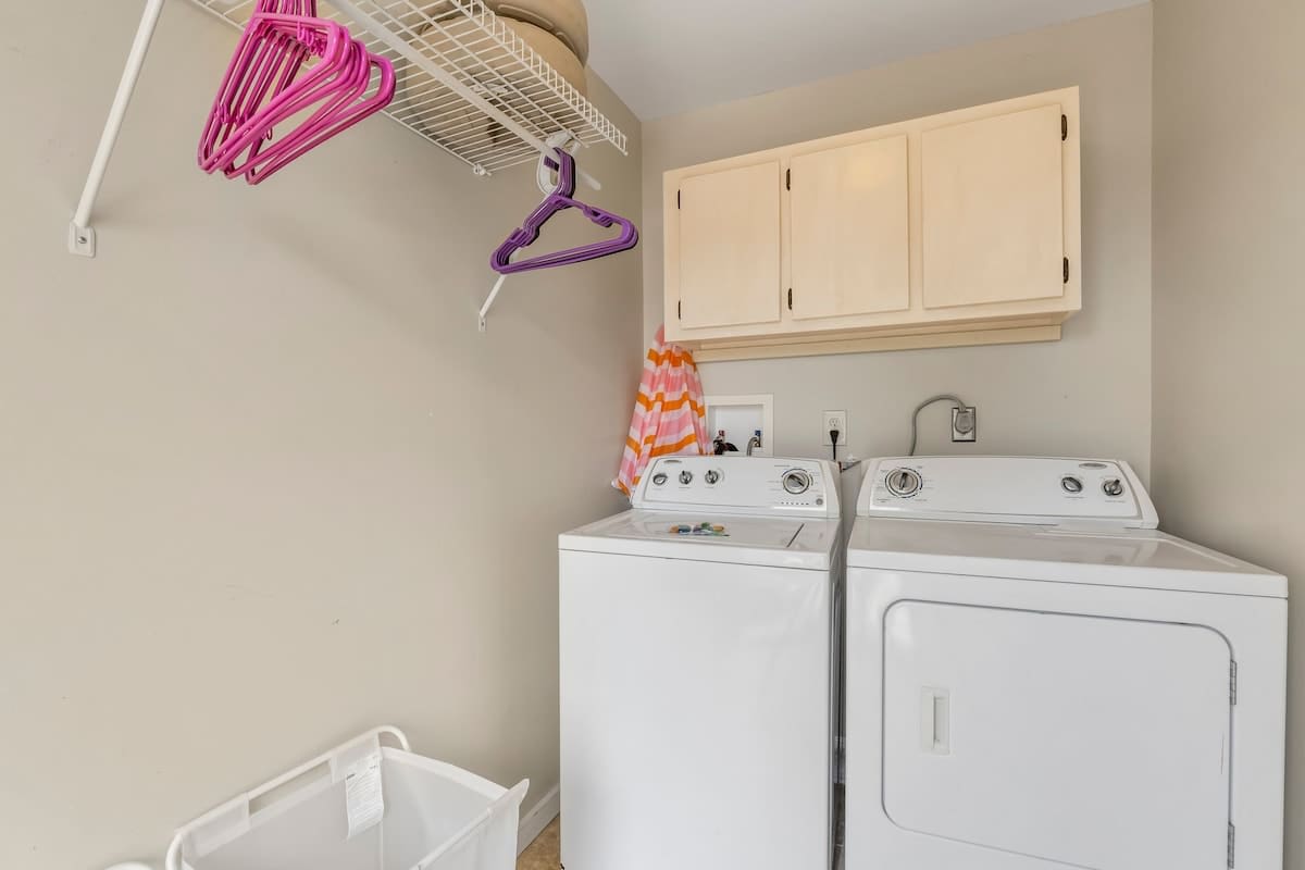 7405 Beach Dr A – Laundry room with white washing machine and dryer, beige walls, hanging pink and purple clothes hangers.