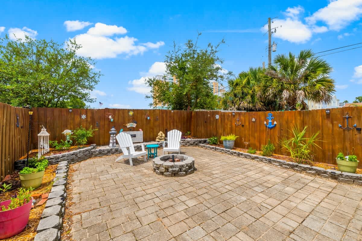 7405 Beach Dr A – Paved outdoor patio with fire pit, two white chairs, and decorative plants surrounded by a wooden fence.