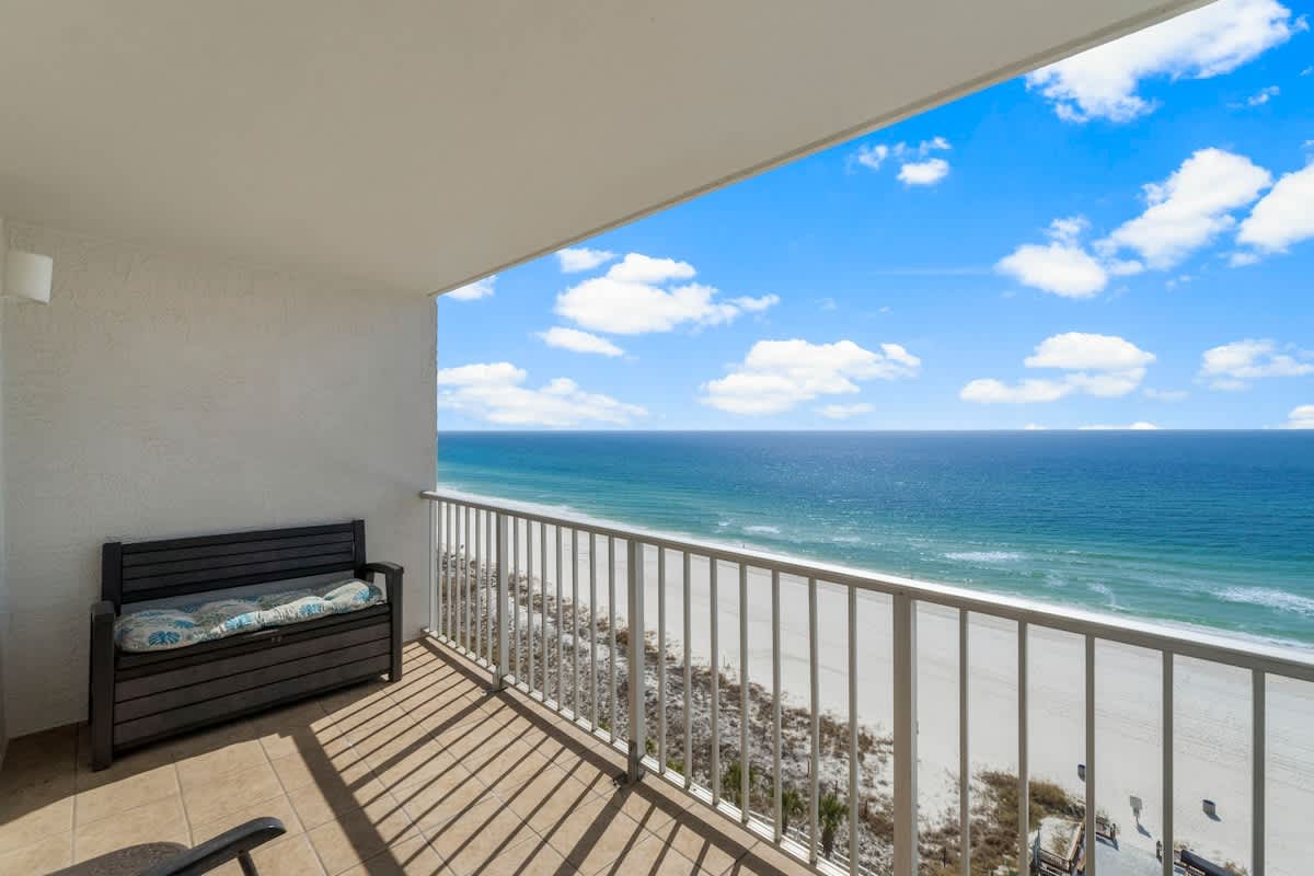 The Summit 1216 – Balcony view of the beach with ocean and blue sky, featuring a bench and tile flooring.