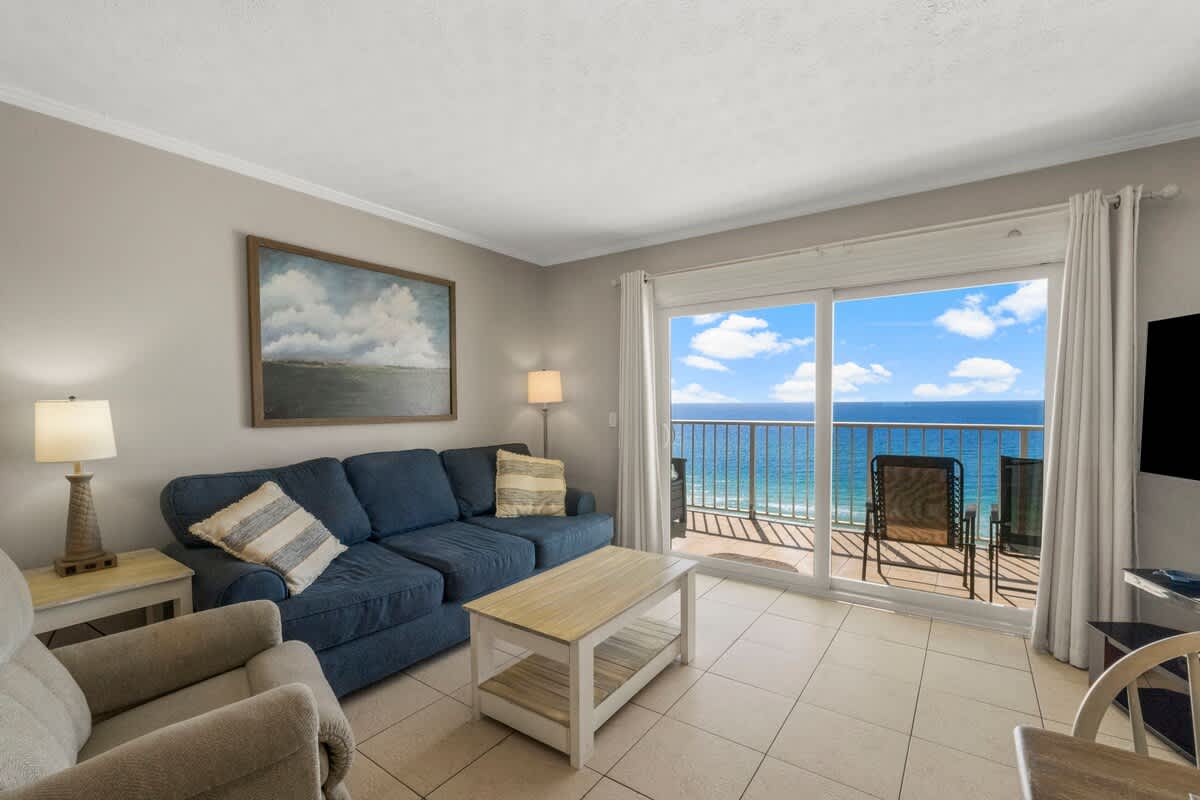 The Summit 1216 – Living room with blue couch, coffee table, beach view, and sliding glass doors to balcony.