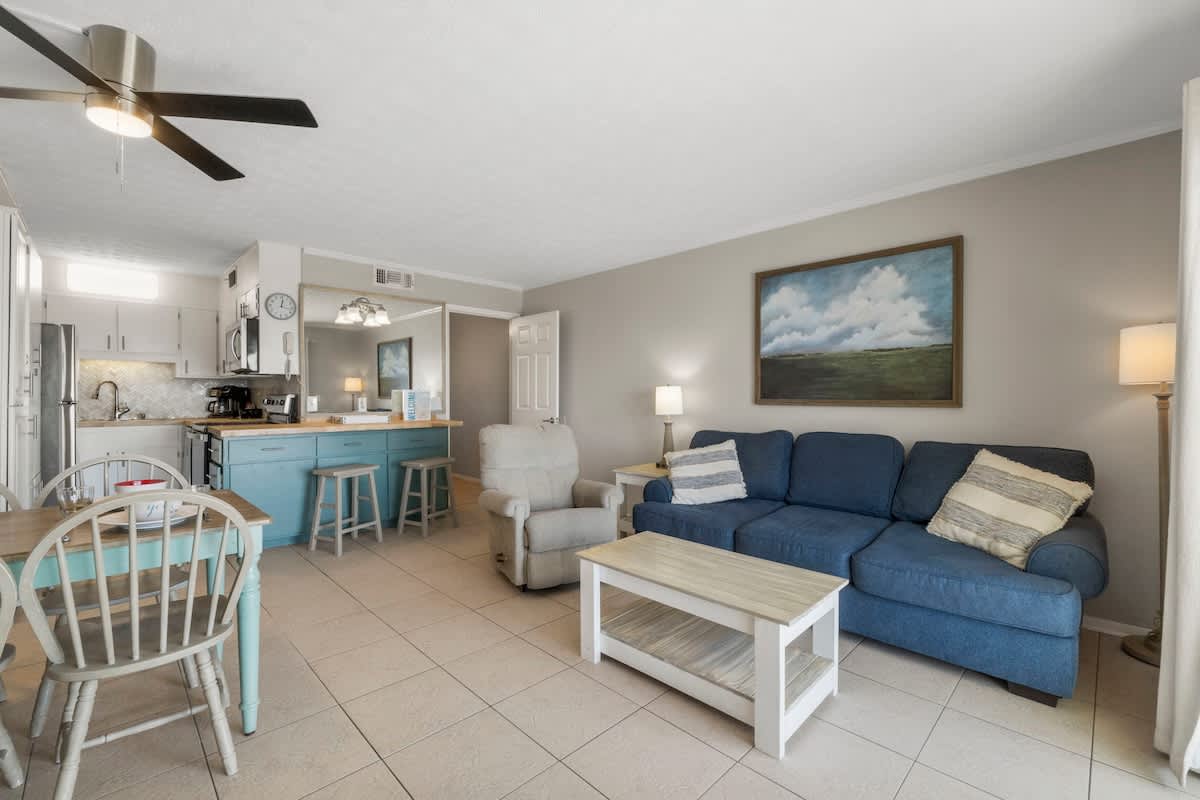 The Summit 1216 – Living room with blue couch, dining table, kitchen area, and natural light from windows.