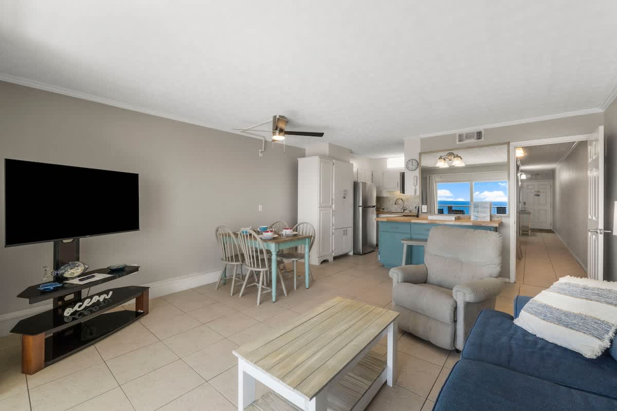 The Summit 1216 – Living room with sofa, dining table, kitchen, television, and large window showing ocean view.