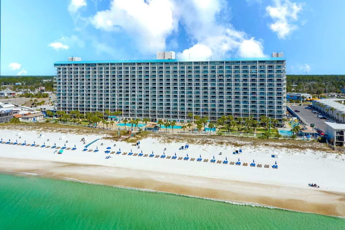 The Summit 1216 – Large beachfront condo building with multiple floors, pool area, and beach chairs on white sand.