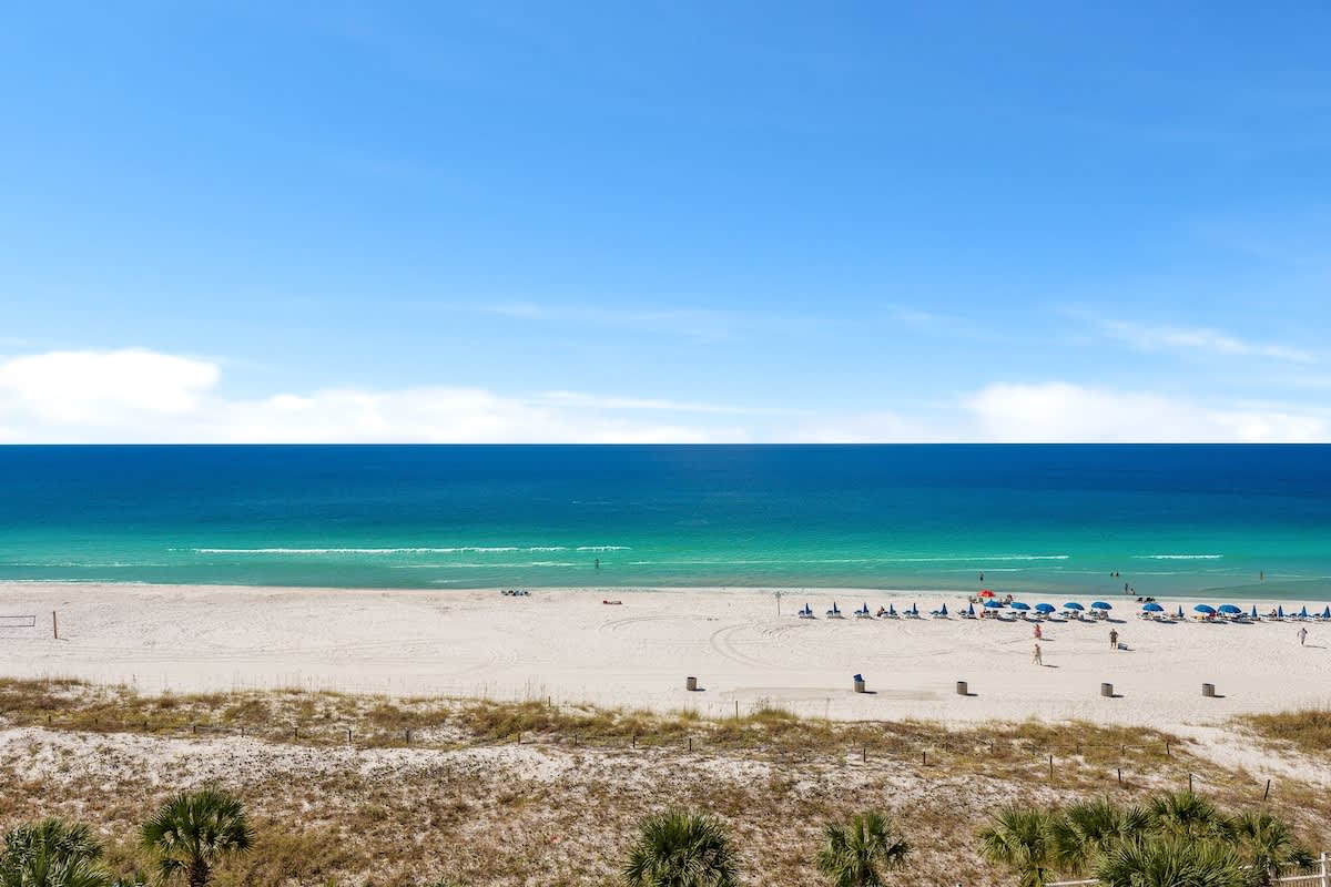 The Summit 726 – Beachfront view with sandy shore, blue umbrellas, and turquoise water under a clear sky.