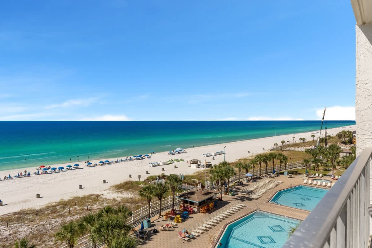 The Summit 726 – Balcony view of two pools, palm trees, and a beachfront with umbrellas and lounge chairs.