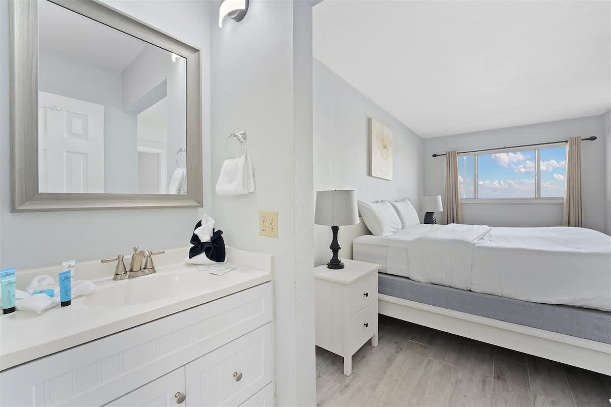 The Summit 1006 – Bathroom with sink and mirror beside a bedroom featuring a bed and window with a cloud view.