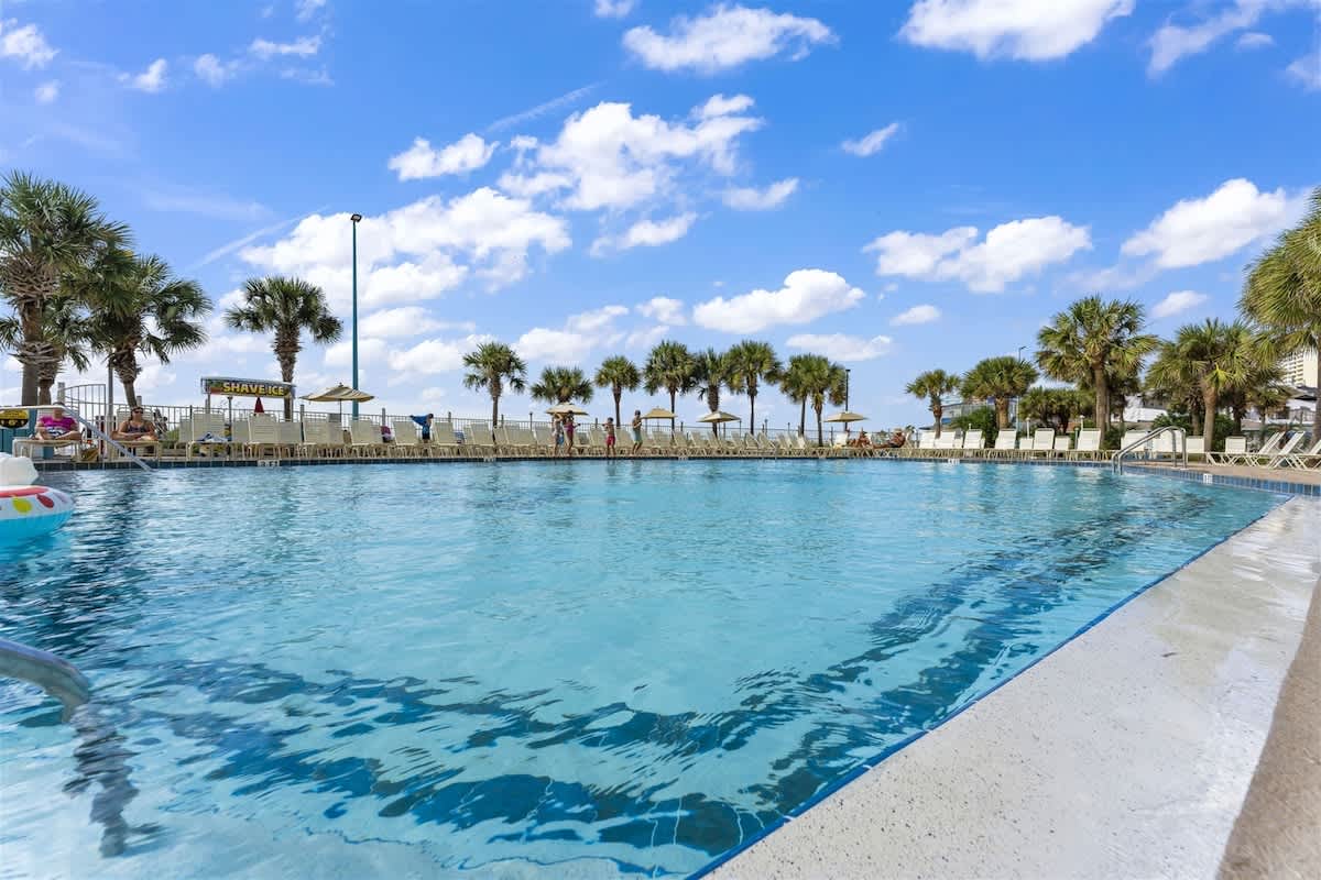 The Summit 1006 – Outdoor swimming pool surrounded by palm trees and lounge chairs under a blue sky with clouds.