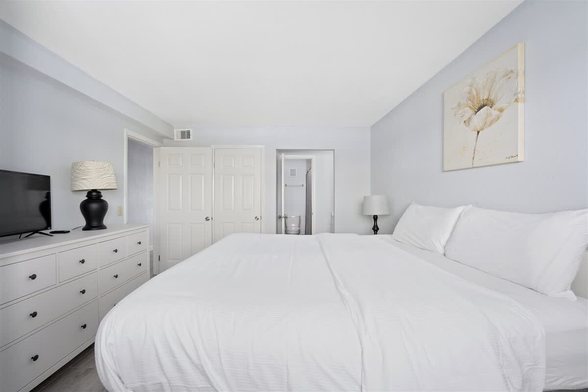 The Summit 1006 – Bedroom with a large bed, dresser, TV, lamp, and an adjacent bathroom door.