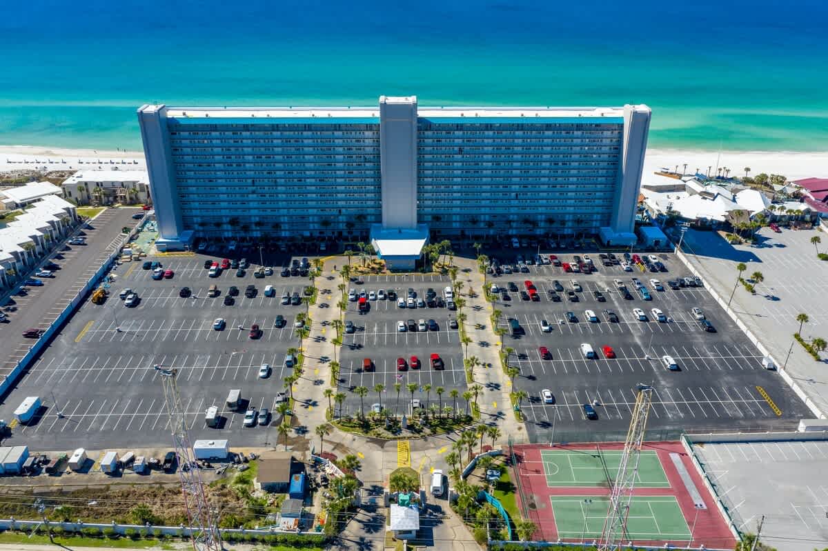 The Summit 1031 – Aerial view of a tall hotel building, parking lot, and nearby beach with turquoise waters.
