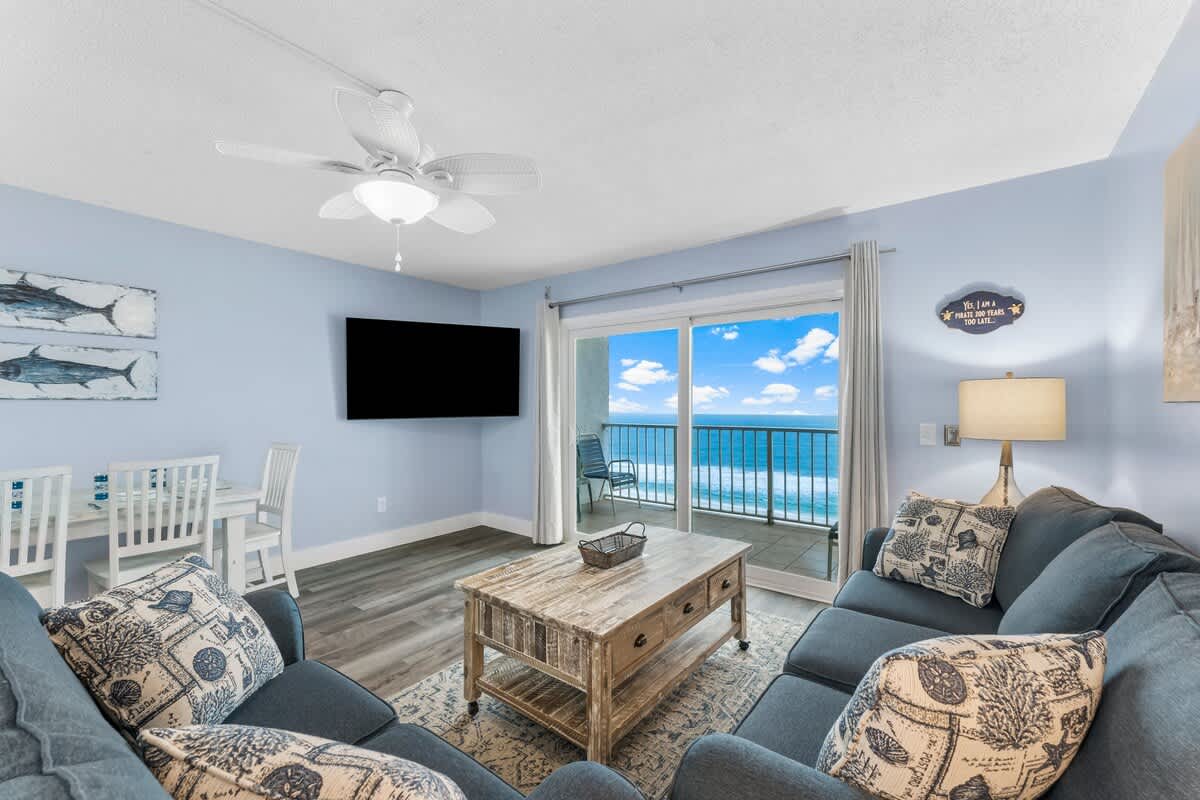 The Summit 1031 – Living room with blue walls, couch, wooden coffee table, dining table, flat-screen TV, and ocean view.