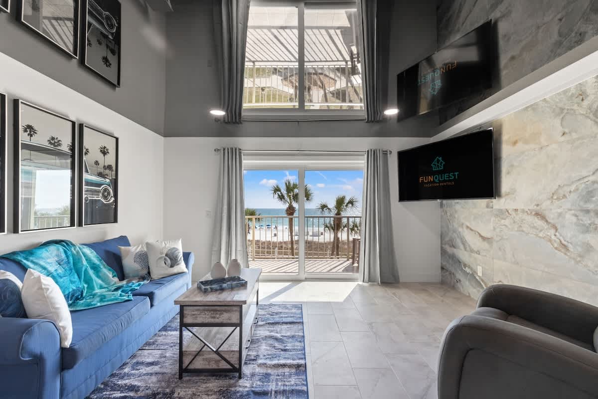 The Summit 306 – Living room with blue sofa, beach view, large windows, and two wall-mounted TVs.