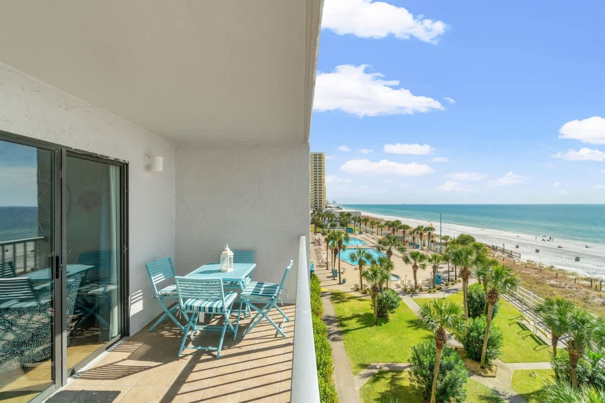 The Summit 502 – Balcony with blue furniture overlooking a beach, pools, and palm trees.