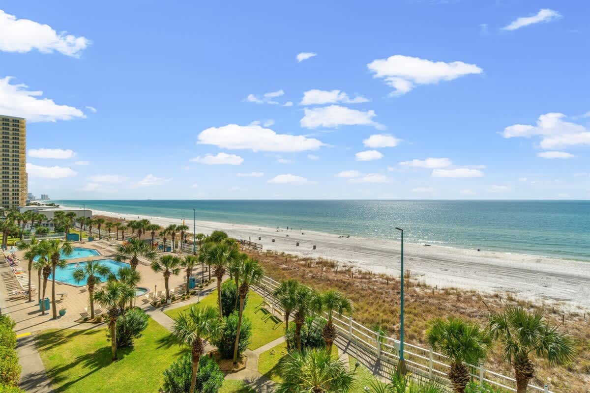 The Summit 502 – Ocean view from a balcony overlooking a pool and sandy beach with palm trees.