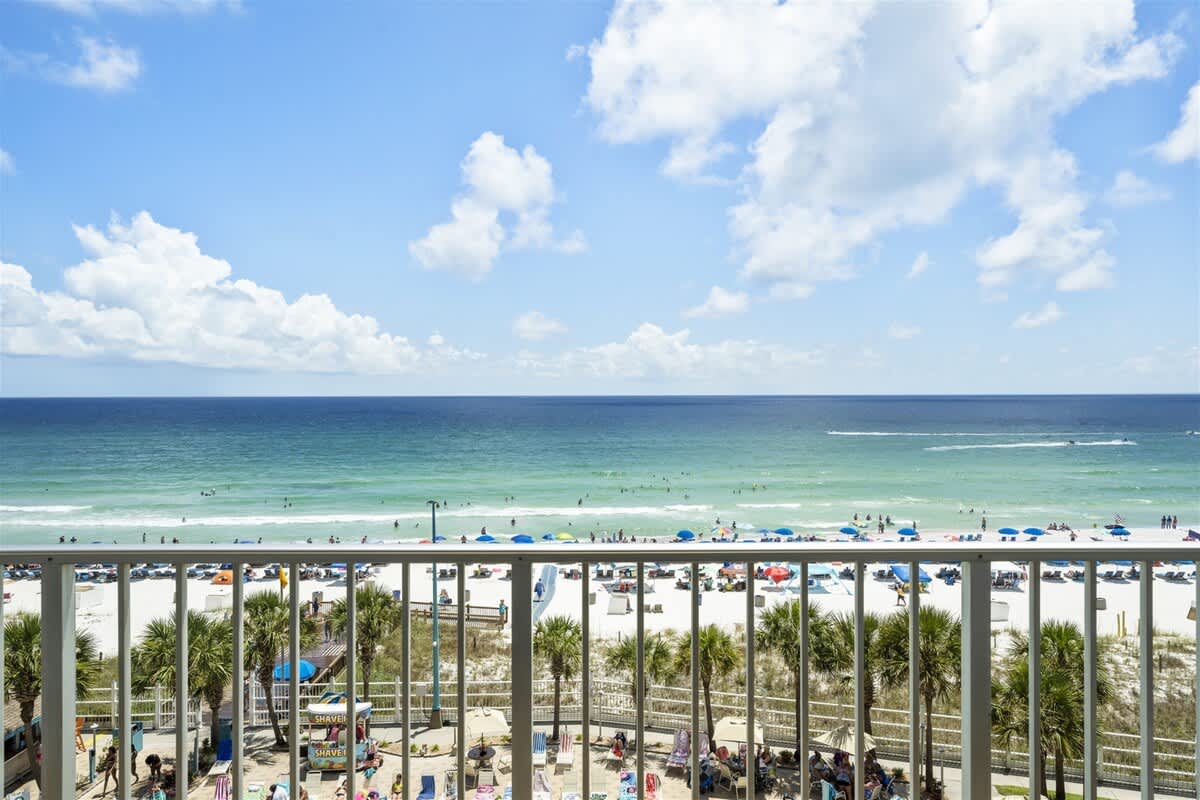 The Summit 613 – Balcony view of a beach with umbrellas, waves, and palm trees under a clear sky.