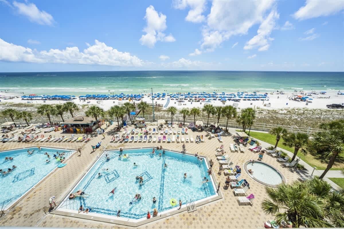 The Summit 613 – Poolside view with two swimming pools, palm trees, and beach with umbrellas in the background.