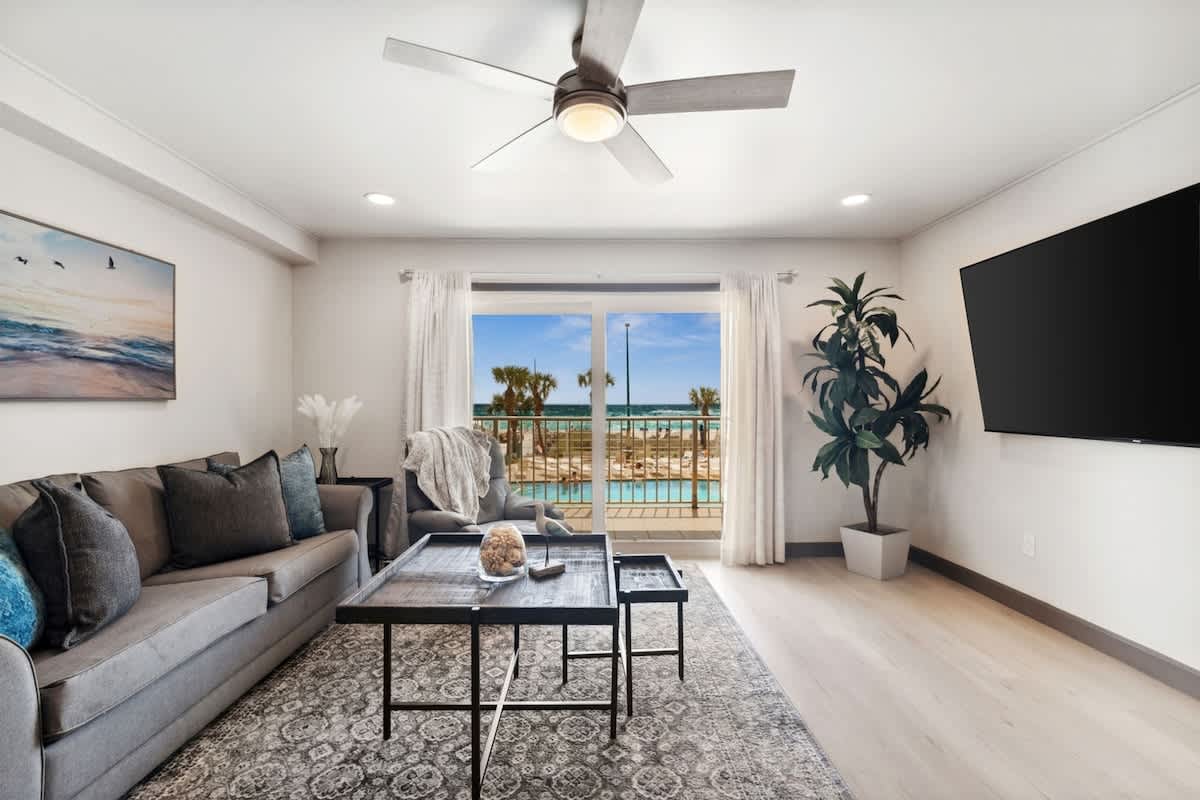 The Summit 215 – Living room with gray sofa, coffee table, TV, indoor plants, and balcony view of palm trees and pool.