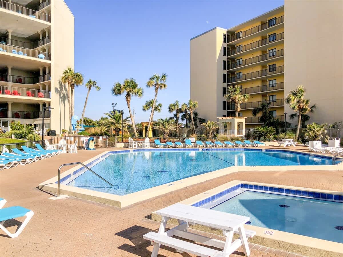 Top Of The Gulf 607 – Pool area with lounge chairs, palm trees, and adjacent building visible in bright sunlight.
