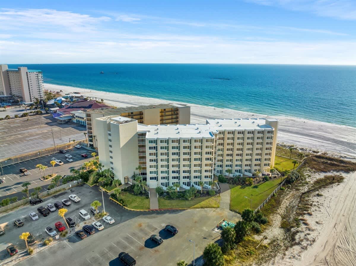 Top Of The Gulf 607 – Aerial view of a beachfront vacation rental building with parking lot and ocean in the background.