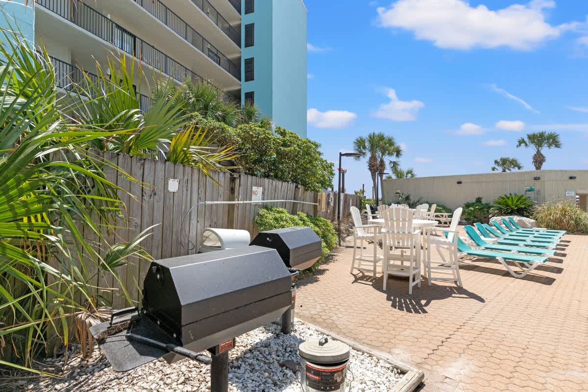 Top Of The Gulf 607 – Outdoor area with grills, seating, and lounge chairs surrounded by landscaping and palm trees.