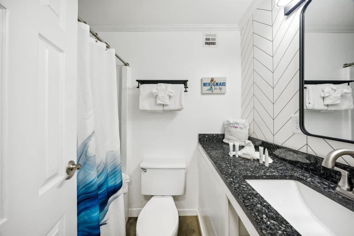 Top Of The Gulf 607 – Bright bathroom with a dark granite countertop, white cabinetry, and a blue wave shower curtain.