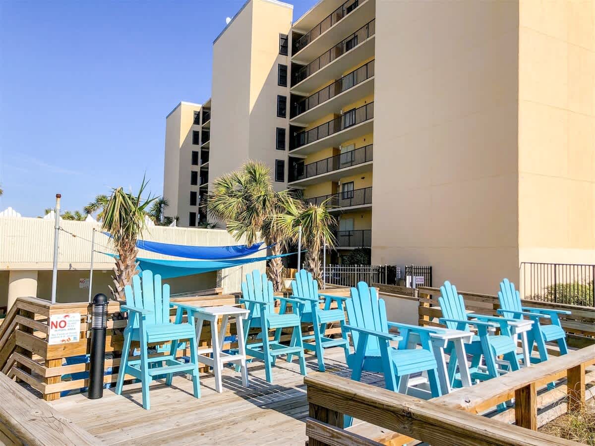 Top Of The Gulf 607 – Wooden deck with blue chairs, palm trees, and a hammock near a tall building in sunny weather.