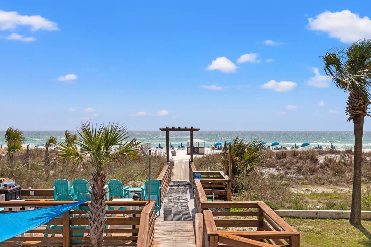Top Of The Gulf 607 – Wooden walkway leading to the beach, with palm trees and lounge chairs visible.
