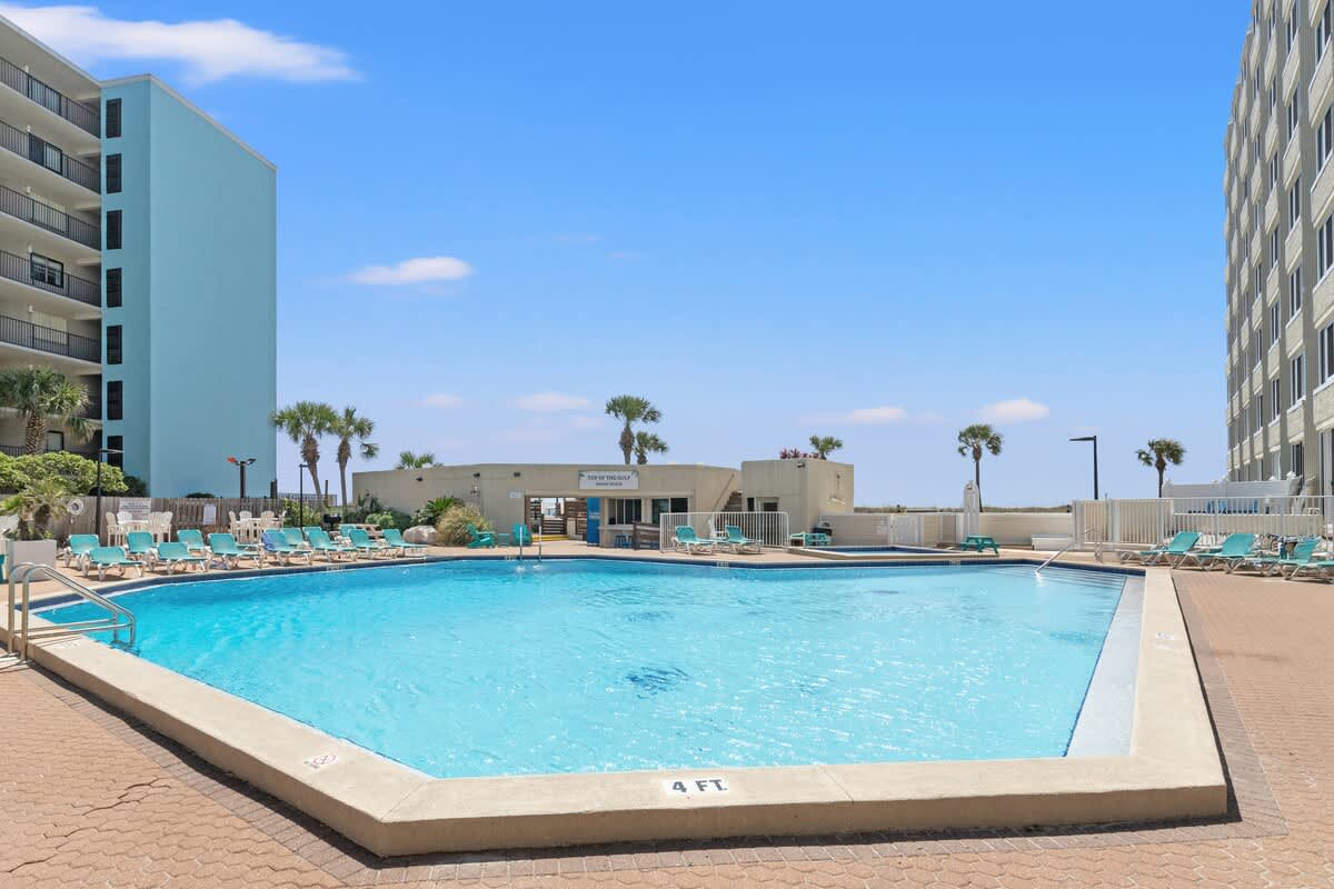 Top Of The Gulf 607 – Swimming pool area with lounge chairs, surrounded by buildings and palm trees under a clear blue sky.