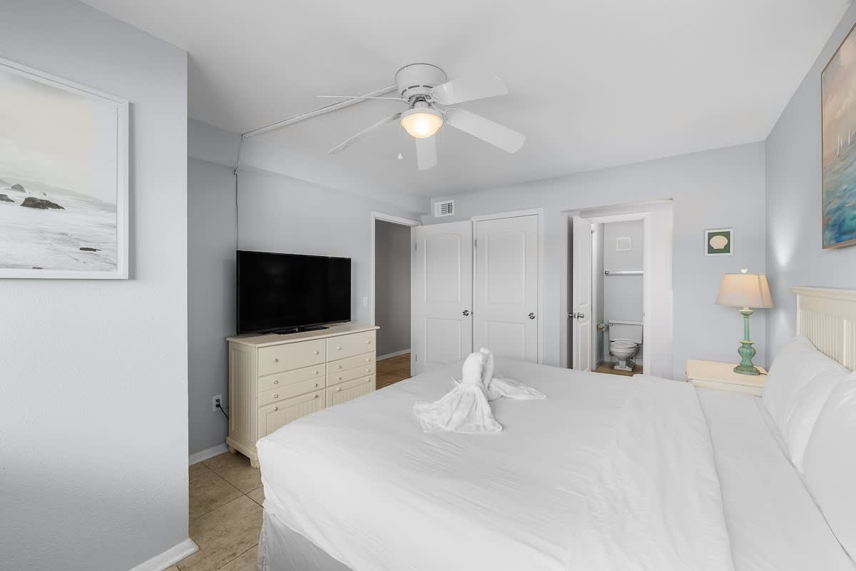 The Summit 606 – Bedroom with king bed, television, dresser, and bathroom entrance; light-colored walls and decorative artwork.