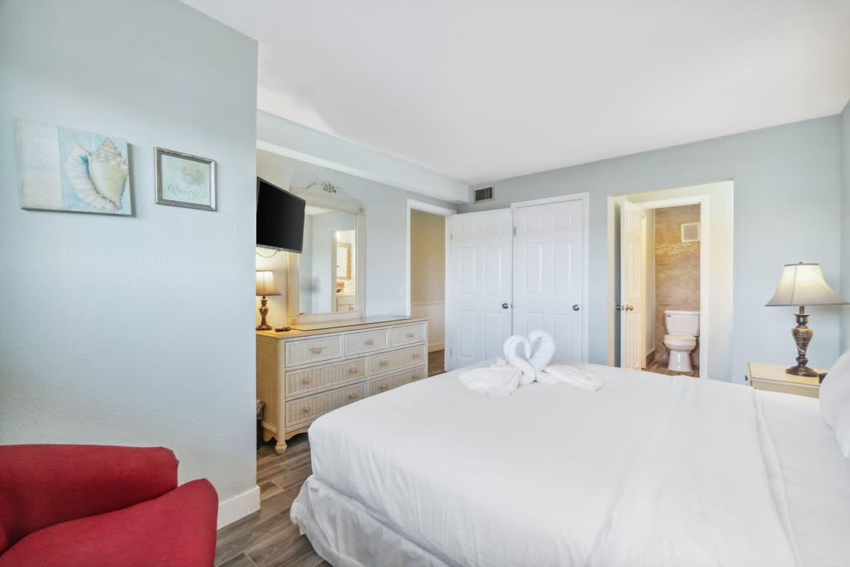 The Summit 108 – Bedroom with white bed, dresser, wall-mounted TV, and bathroom doorway in the background.
