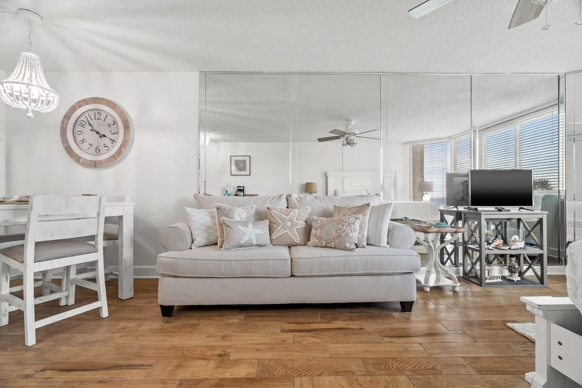 Top Of The Gulf 213 – Living room with a beige sofa, dining area, wooden flooring, and large windows with blinds.